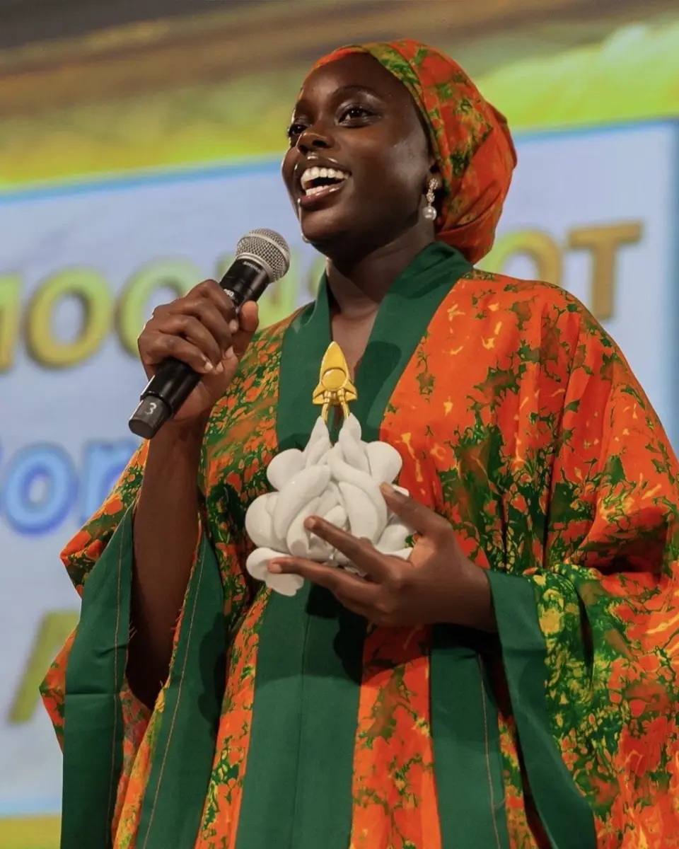 Denise Ayebare on stage, speaking into a microphone and holding the Moonshot Borderless Award