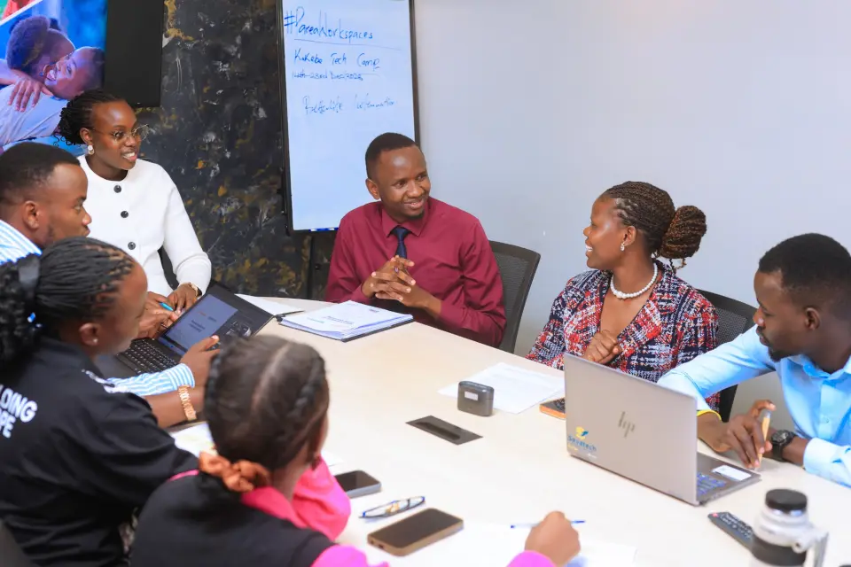 BetterLife and Kakebe representatives talking around a meeting table, beside a whiteboard that reads Kakebe Tech Camp, 14th to 23rd December